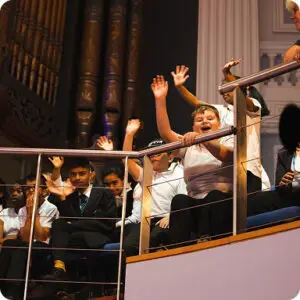 Excited group of students waving from a balcony during a school event in a large auditorium.
