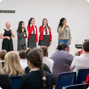 Students perform in front of an audience with a teacher guiding them on stage.