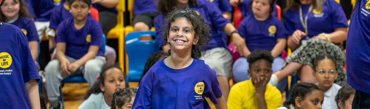 Smiling child in a Music for Life T-shirt at an event with other children wearing matching shirts in the background.