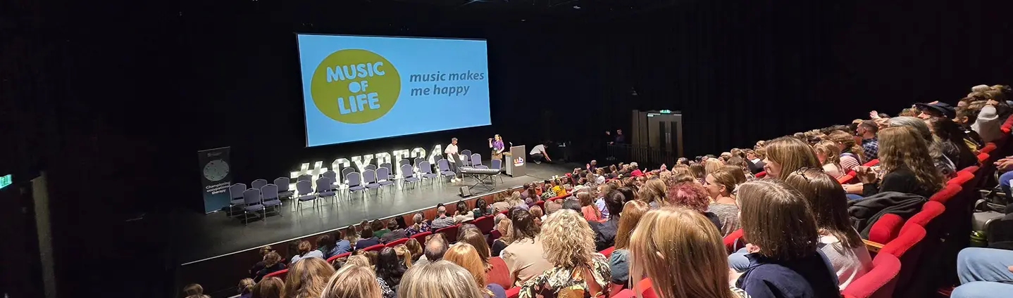 Audience at a Music of Life event in a theater setting, with a screen displaying the slogan Music makes me happy.