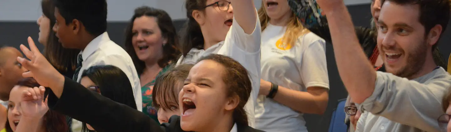 Diverse group of people enthusiastically singing and cheering at an event, raising their arms in excitement.