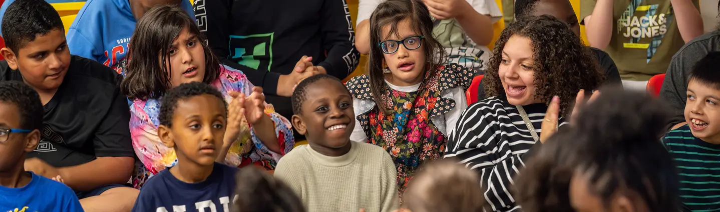Diverse group of children sitting together, smiling and clapping at a school event, creating a joyful atmosphere.