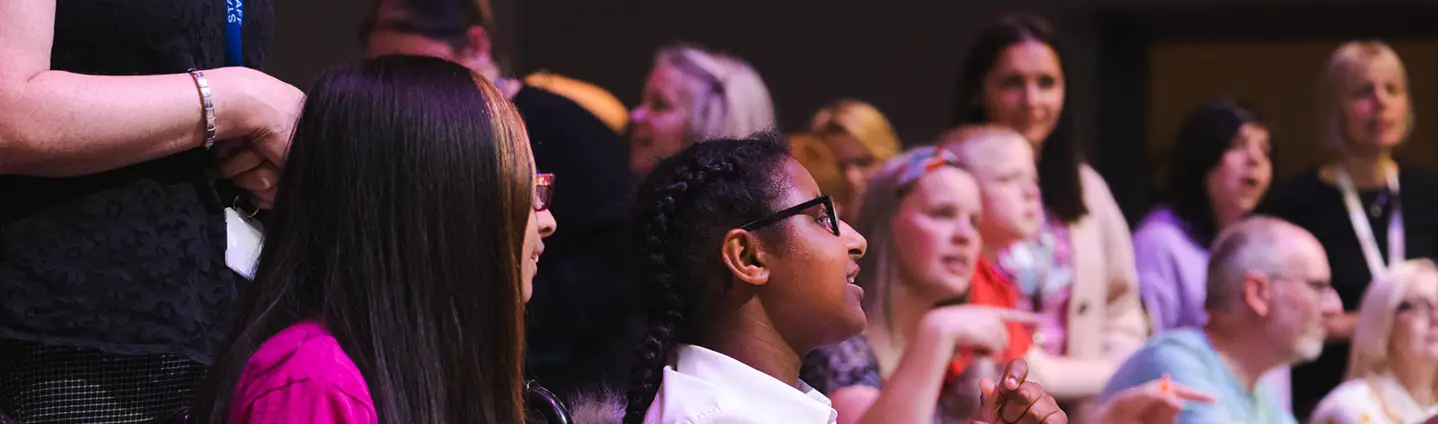 Audience watching a live event, diverse crowd engaged and smiling, focus on two people in the foreground.