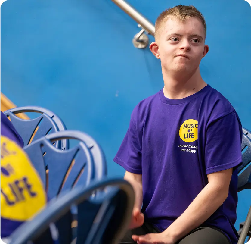 Young man in a purple Music of Life shirt smiles while sitting on a chair, enjoying a musical event.