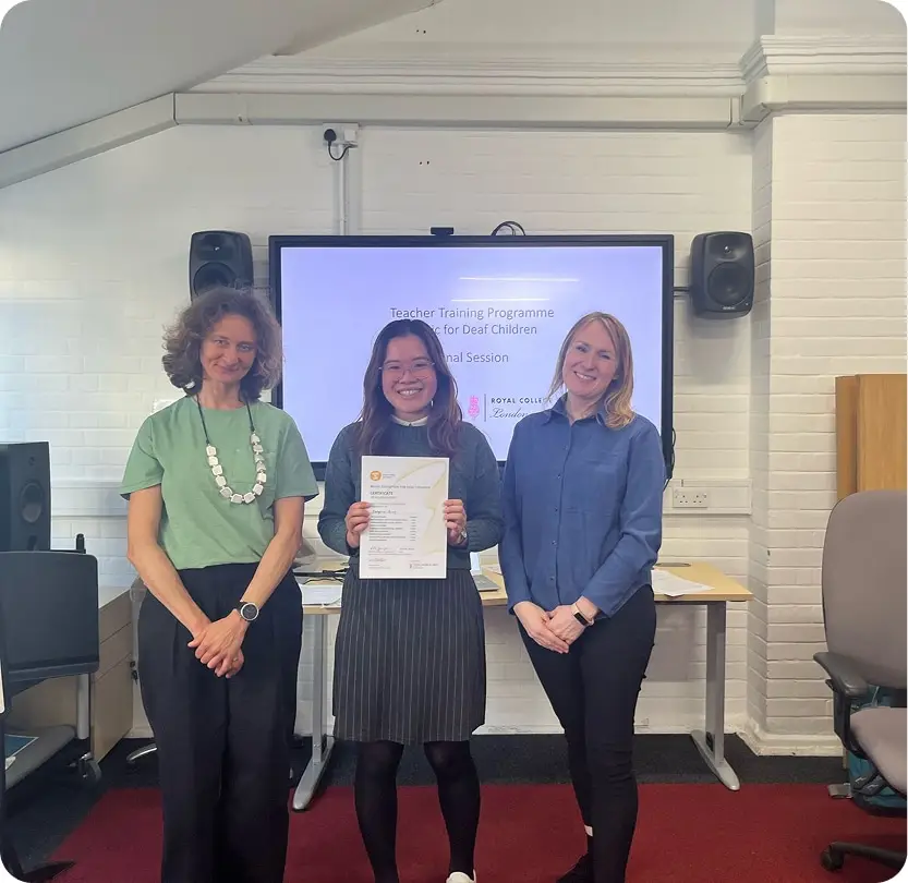 Three people at a teacher training program for music and deaf children, one holding a certificate, smiling.