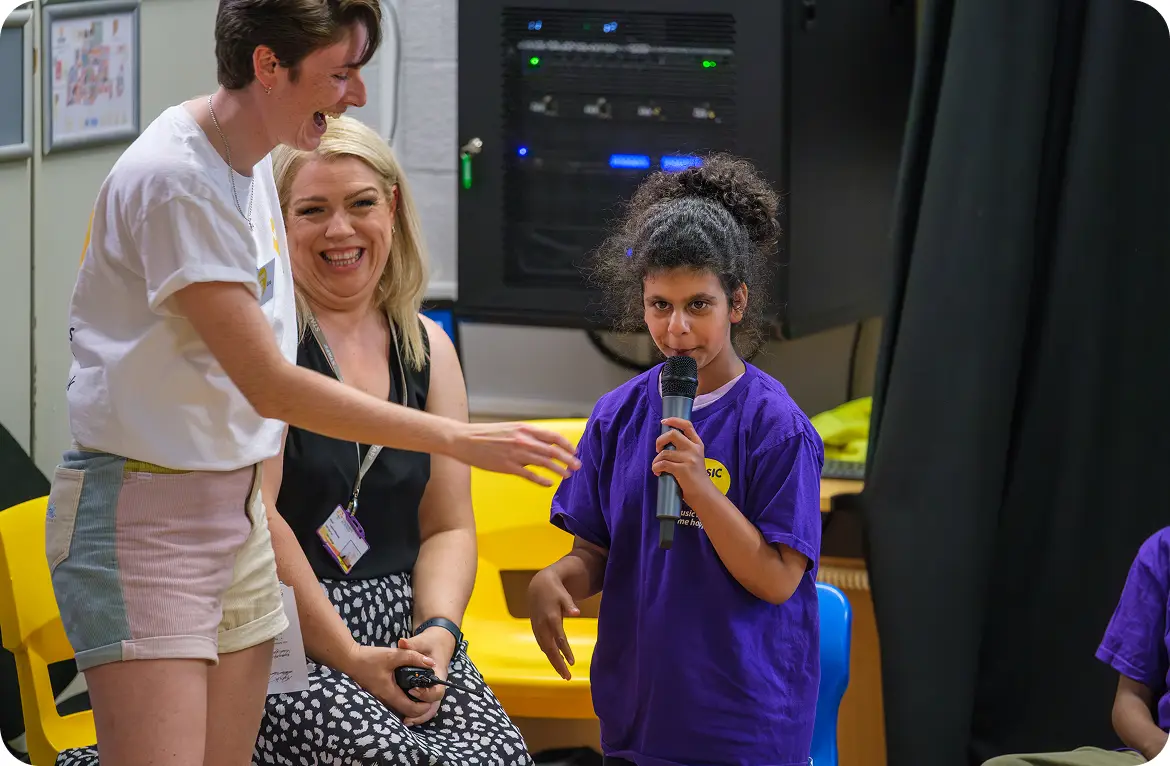 Child speaking into a microphone at an event with two women smiling in the background.