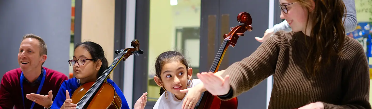 A diverse group of children learning music together, playing cellos with guidance in a classroom setting.