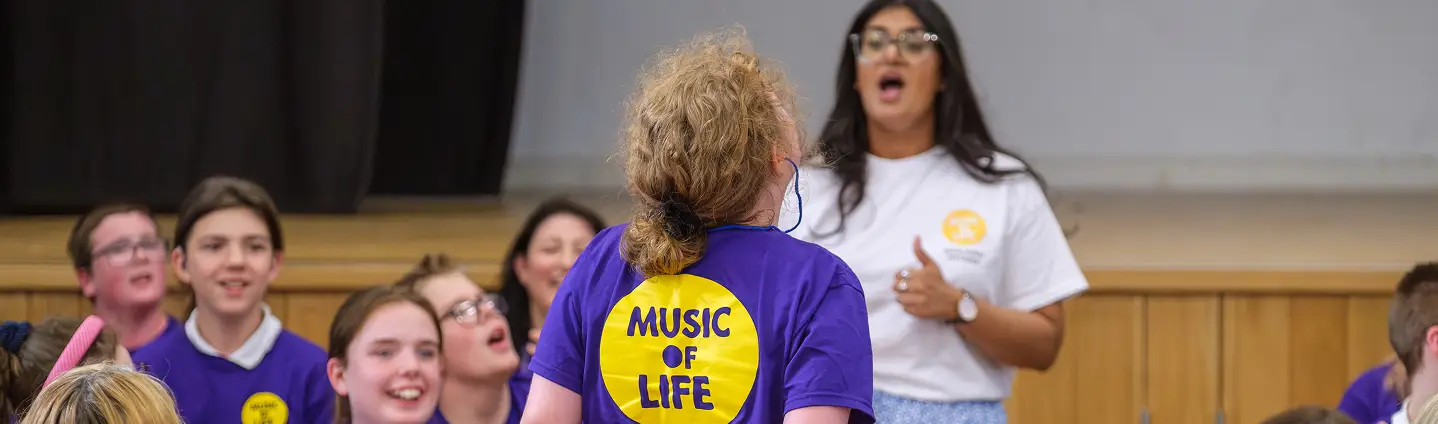 Group of kids in a music program called Music of Life, singing and wearing purple shirts in a classroom setting.