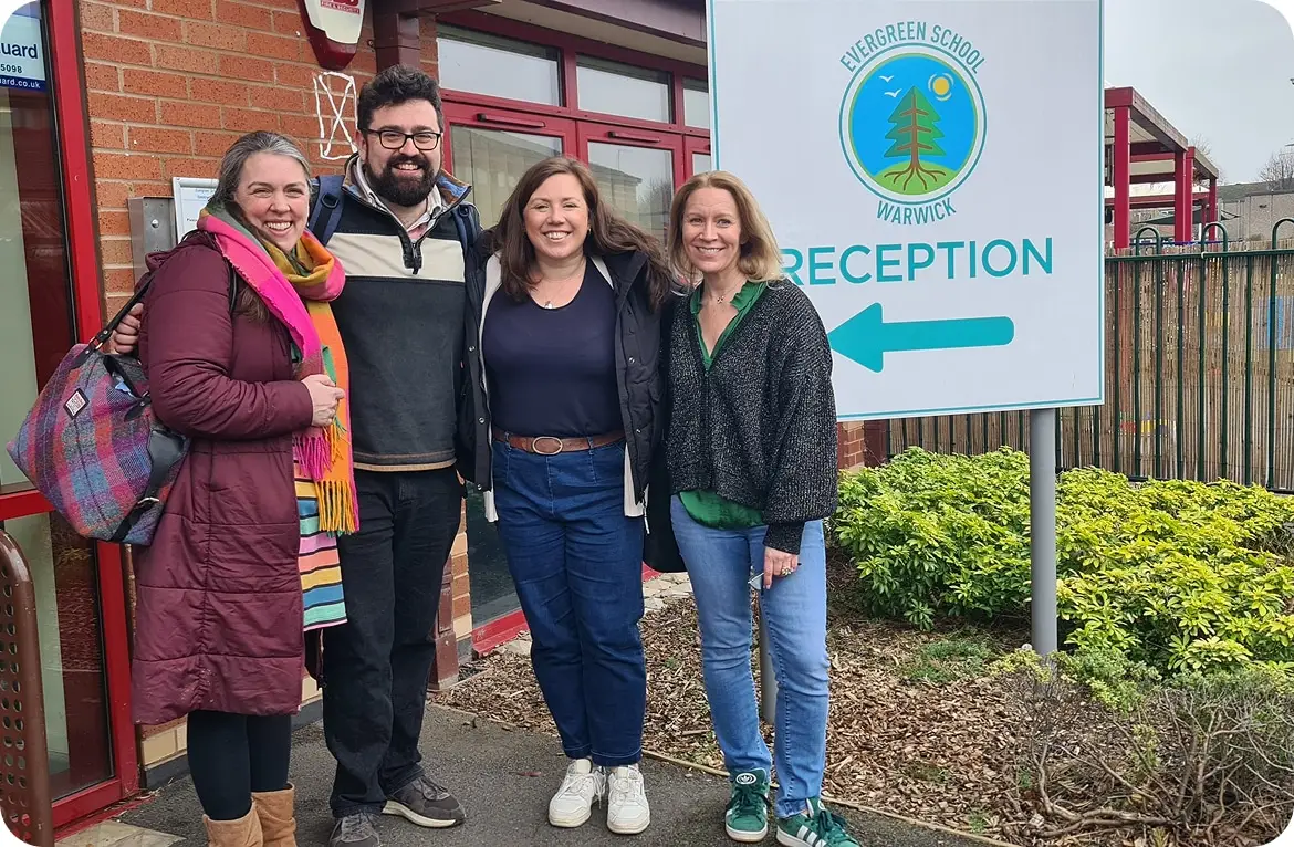 Group of four adults smiling in front of Evergreen School Warwick reception sign.