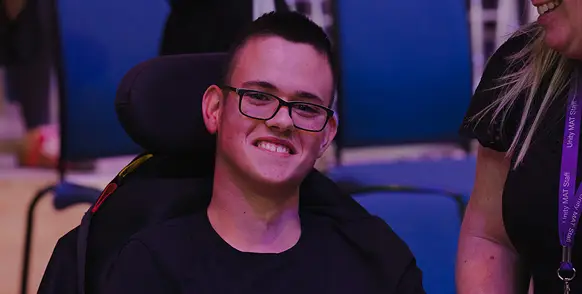 Smiling young man in a wheelchair at an event, wearing glasses and a black shirt, seated in a blue auditorium.