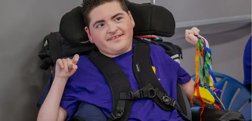 Smiling young person in a wheelchair holding colorful ribbons, wearing a blue shirt, in a cheerful setting.