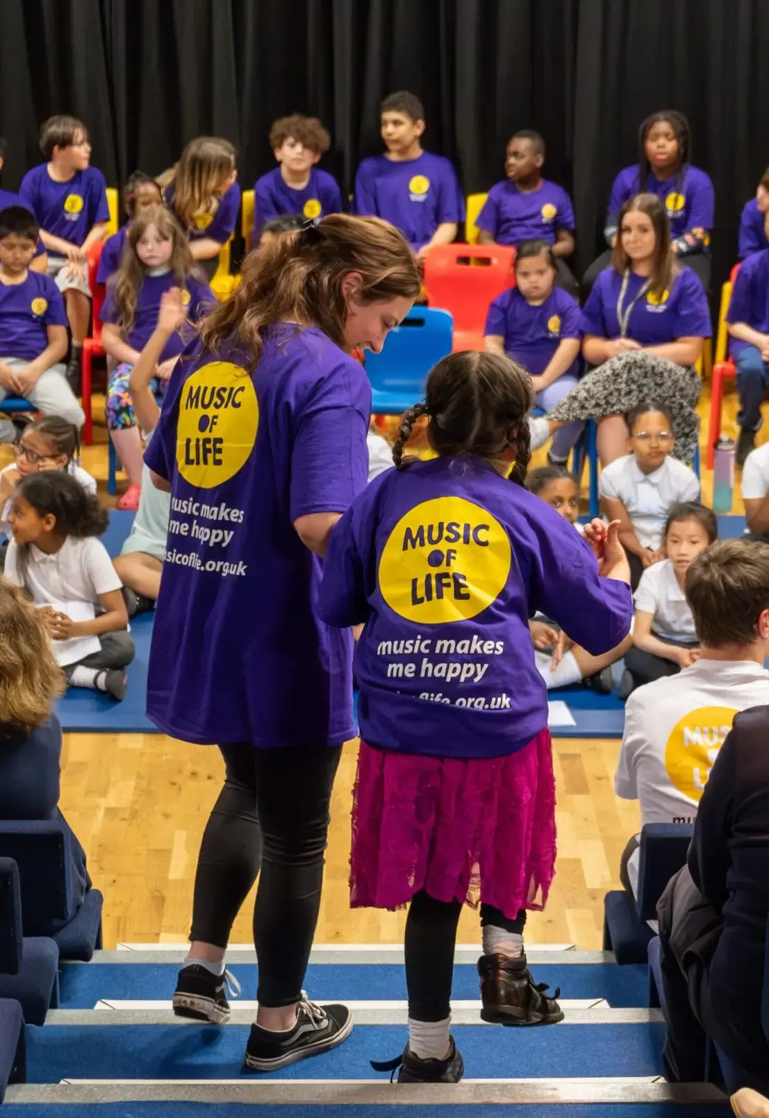 Children in purple Music of Life shirts in a music class setting, enjoying activities and learning together.