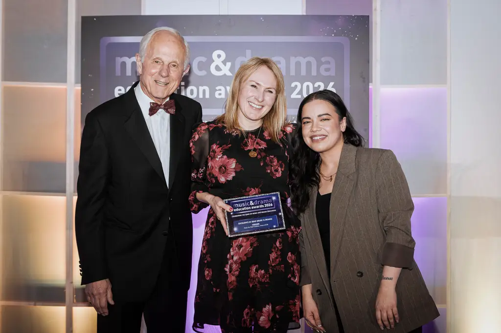 Three people smiling at the Music & Drama Education Awards 2024, one holding a plaque. Elegant setting with soft lighting.