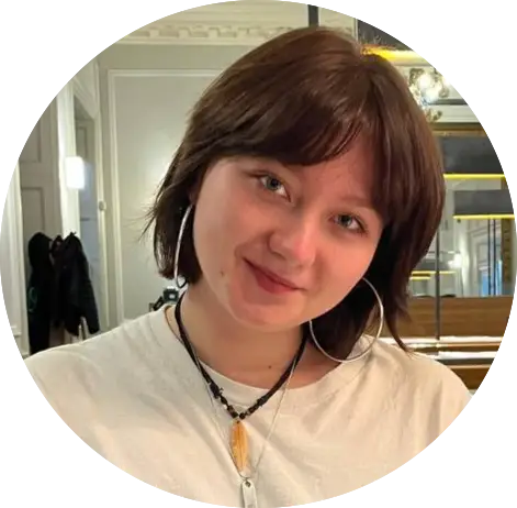 Manya Smiling person with short brown hair, wearing a white shirt and hoop earrings indoors.