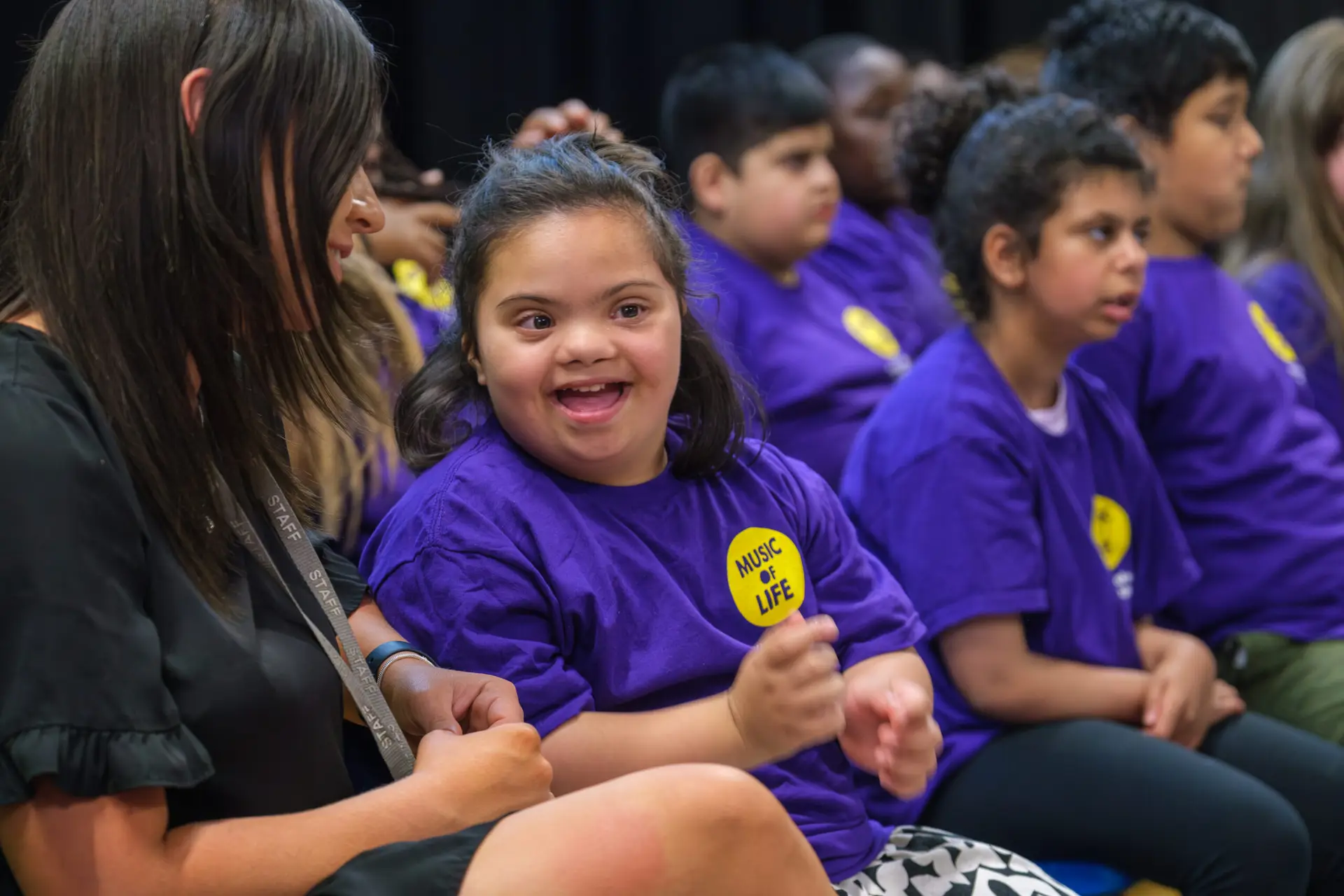 Child smiling in music class, wearing a Music of Life shirt, surrounded by peers and an attentive teacher.