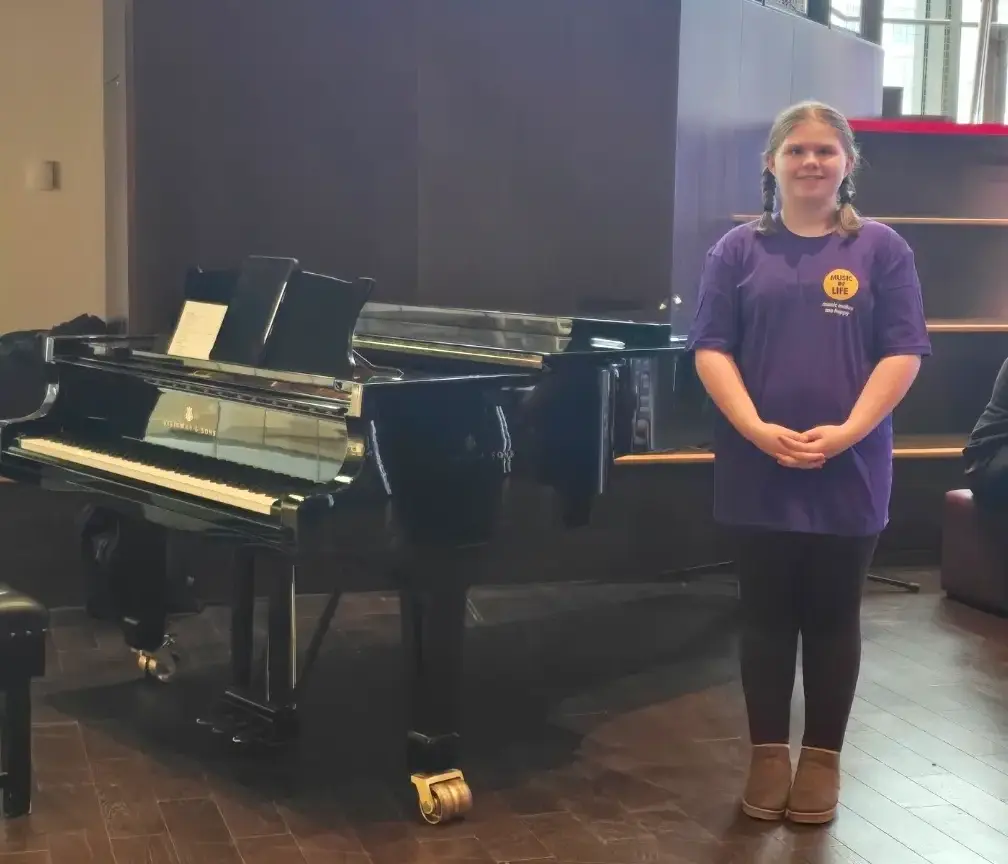Girl in purple shirt stands beside a grand piano in a modern room, showcasing musical talent ambiance.
