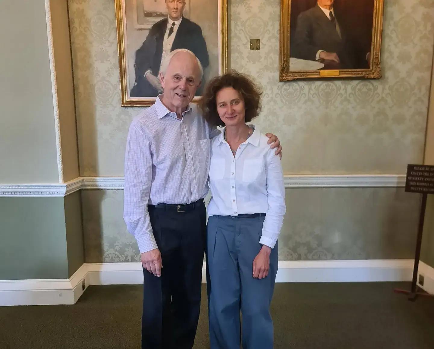 Elderly man and woman smiling in a classic room with portraits on the wall.